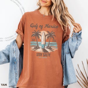May include: A rust-colored short-sleeve t-shirt featuring a graphic of a surfboard, palm trees, and the ocean. The text reads Gulf of Mexico, EST 1550, STAY SALTY, AND RESIST. The shirt is worn under a light blue denim jacket with metal buttons.