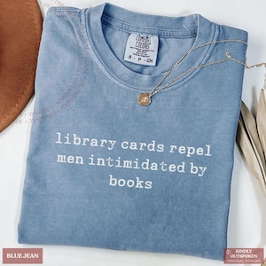 Puede incluir: Una camiseta azul vaquero con el texto blanco "library cards repel men intimidated by books". Un collar dorado con un pequeño colgante descansa sobre la camiseta. La camiseta es de la marca Comfort Colors.