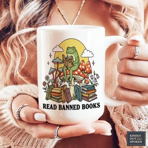 May include: White ceramic mug featuring a whimsical design with a green frog reading a book titled "Banned Tales" atop a mushroom. The mug has the text "READ BANNED BOOKS" and is surrounded by colorful mushrooms and books.