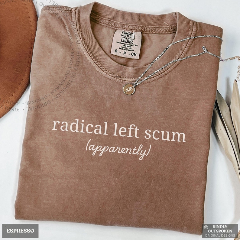 May include: A brown short-sleeve t-shirt with a crew neckline featuring the white printed text radical left scum (apparently) on the front. The shirt has a Comfort Colors brand label inside the collar and is paired with a thin silver-tone necklace.