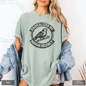 May include: A sage green short-sleeve t-shirt featuring a black circular graphic of a bird perched on a branch. The text "RADICALIZED BY HUMAN DECENCY" is printed in a bold, distressed font around the perimeter of the circular design.
