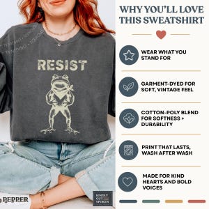 Op de afbeelding: Donkergrijze sweatshirt met het woord "RESIST" boven een kikkerillustratie met een bandana. De sweatshirt heeft een vintage uitstraling. De tekst "WHY YOU'LL LOVE THIS SWEATSHIRT" is ook zichtbaar.