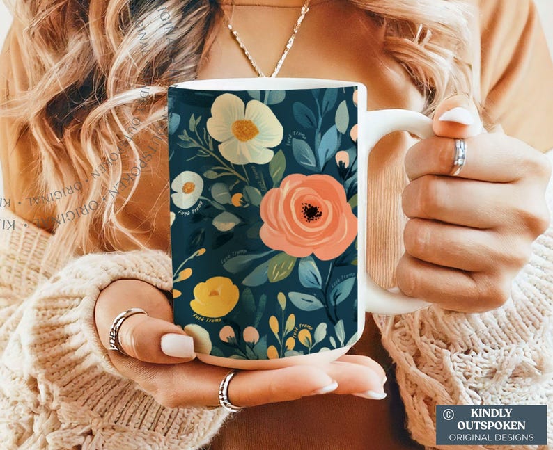 May include: A white ceramic mug featuring a dark teal background with a floral pattern of large peach, cream, and yellow flowers surrounded by green and blue leaves. The text "KINDLY OUTSPOKEN ORIGINAL DESIGNS" is printed in a small box at the bottom.