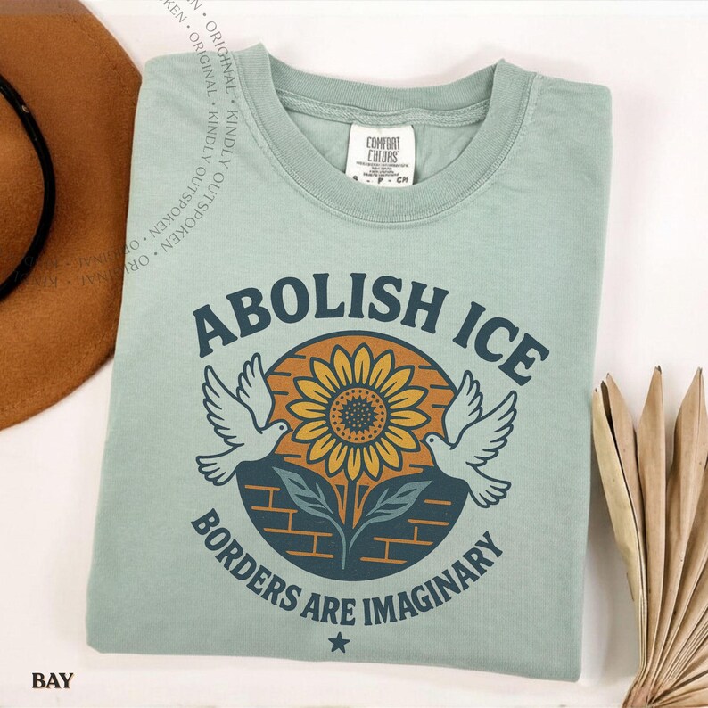 May include: A sage green t-shirt featuring a graphic print of a yellow sunflower centered within a dark blue circle. Two white doves flank the flower. The text reads ABOLISH ICE above and BORDERS ARE IMAGINARY below, with a small star at the bottom.