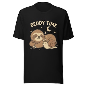 Puede incluir: Camiseta negra con un perezoso y un caracol de dibujos animados durmiendo bajo una luna creciente y estrellas. El texto "BEDDY TIME" está encima de la ilustración. El diseño está impreso en una camiseta suave y cómoda.