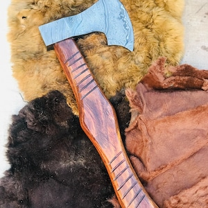 May include: A handcrafted axe with a patterned steel blade and a polished wooden handle. The handle features carved grooves for grip. The axe is displayed on a textured, brown fur background, showcasing its rustic design.