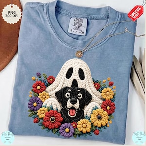 May include: A light blue Comfort Colors t-shirt featuring a graphic of a black dog peeking out from behind a white ghost, surrounded by colorful flowers. A silver necklace with a pendant is draped across the shirt. The shirt has the text "COMFORT COLORS" on the tag.
