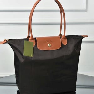 May include: A black nylon tote bag featuring tan leather handles and a matching tan leather flap closure with a gold-tone snap button. The bag includes a green rectangular tag attached to the side and a gold-tone zipper pull on the top opening.