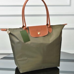 May include: An olive green tote bag featuring a brown leather flap with a gold-tone snap button closure, two matching brown leather handles, and a small green rectangular tag attached to the side near the top zipper pull.