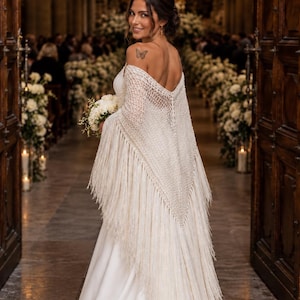 May include: A cream-colored, off-the-shoulder crochet shawl featuring an intricate open-knit pattern and long, flowing fringe along the edges. The shawl is draped over a smooth, floor-length white gown, creating a layered, textured appearance.