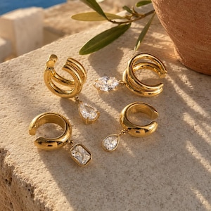 Può includere: Quattro ear cuff color oro con finitura lucida. Due presentano un design a doppio cerchio con un cristallo trasparente a goccia pendente, mentre gli altri due sono a cerchio singolo, uno con un cristallo a goccia e uno con un cristallo rettangolare.