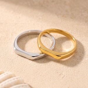 Puede incluir: Dos anillos de metal liso y pulido con un diseño de parte superior plana. Un anillo tiene un acabado en tono plateado brillante, mientras que el otro tiene un acabado en tono dorado cálido. Ambos anillos presentan una banda simple y redondeada.