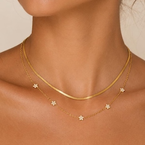May include: A layered gold necklace set consisting of a flat herringbone chain and a delicate cable chain. The lower chain features evenly spaced small star-shaped charms, each embedded with tiny clear stones that catch the light.