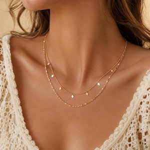 May include: A double-layered gold necklace featuring a delicate beaded chain with small, evenly spaced diamond-shaped charms and a second, slightly longer chain with a twisted link design. The jewellery rests against the neck with a polished finish.