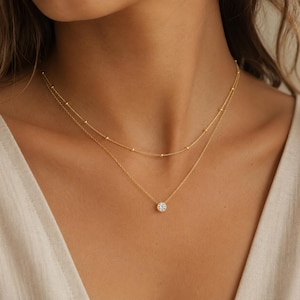 Puede incluir: Un conjunto de collares dorados superpuestos que consta de dos cadenas delicadas. La cadena superior presenta pequeñas cuentas doradas espaciadas uniformemente, mientras que la cadena inferior sostiene un colgante redondo de piedra clara engastada.