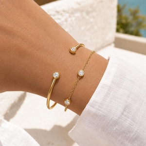 May include: A gold-toned open cuff bracelet featuring two clear round stones at the ends, paired with a delicate gold-toned chain bracelet adorned with three evenly spaced clear round stones, both worn together on the wrist.