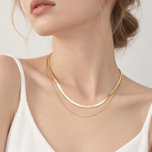 Puede incluir: Un collar de capas en tono dorado que presenta una cadena plana y ancha de espiga junto a una segunda cadena fina y delicada. El acabado metálico dorado refleja la luz, creando una apariencia pulida alrededor del cuello, combinado con una prenda blanca.