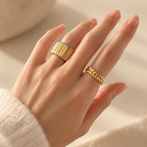 Op de afbeelding: Twee goudkleurige ringen gedragen aan de vingers. De ene ring is een brede band met een verticale ribbeltextuur. De tweede ring is een smallere band met een gevlochten of korenaarachtig patroon. Beide ringen hebben een gepolijste gouden afwerking.