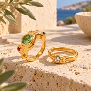 Pode incluir: Dois anéis dourados com bandas irregulares e texturizadas. O anel maior apresenta uma pedra redonda verde proeminente, uma pequena pedra vermelha, uma minúscula pedra amarela e uma pedra azul escura. O anel menor tem uma pedra clara.