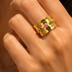 Peut inclure: Une large bague en métal doré avec une surface martelée et texturée. L'anneau est orné de cinq petites pierres précieuses rondes et multicolores disposées en ligne horizontale, incluant des pierres roses, bleues, vertes et transparentes.