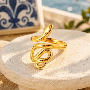 May include: A gold-toned ring shaped like a coiled snake. The metal has a polished, reflective surface. The snake's body forms the band of the ring, with the head extending upwards and the tail curving to complete the circular design of the jewellery piece.