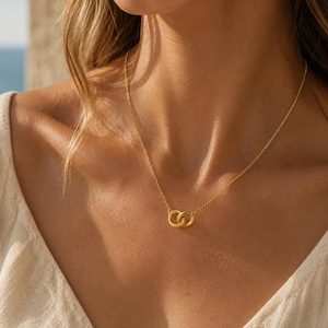 Puede incluir: Un collar de tono dorado que presenta dos pequeños anillos circulares entrelazados como colgante central. El colgante está suspendido de una delicada y fina cadena de tono dorado que descansa sobre la clavícula, mostrando un acabado metálico pulido.