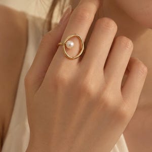 Puede incluir: Un anillo de tono dorado con una banda circular abierta e irregular y una perla blanca única colocada de forma descentrada en el espacio abierto. El metal tiene un acabado liso y pulido, creando un diseño delicado y de marco abierto.