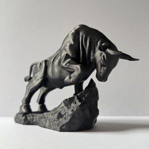 May include: A dark grey sculpture of a bull in a charging pose, mounted on a textured rock base. The bull's muscular form is detailed, with its head lowered and horns forward, conveying a sense of power and determination. The sculpture is likely made of a durable material.