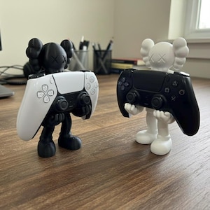 May include: Two figurines, one black and one white, each holding a PlayStation 5 controller. The black figurine has a black body and a white controller. The white figurine has a white body and a black controller.