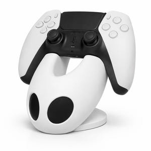 Könnte beinhalten: Ein weiß-schwarzer Gamecontroller, der auf einem passenden weißen Ständer ruht. Der Controller hat einen schwarzen Mittelteil mit Knöpfen und Joysticks sowie weiße Seitengriffe. Der Ständer hat ein einzigartiges, geschwungenes Design mit zwei großen, kreisförmigen Öffnungen.
