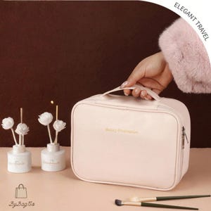 Può includere: Un beauty case da viaggio rosa chiaro con manico, cerniera e la scritta "Beauty Preservation" in lettere dorate. Due pennelli per il trucco e due piccoli diffusori di fiori bianchi in primo piano.