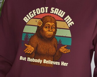 Puede incluir: Sudadera con capucha burdeos con un gráfico de Bigfoot. El gráfico presenta un Bigfoot de dibujos animados con una flor, sobre un fondo de puesta de sol. El texto dice "BIGFOOT SAW ME" y "But Nobody Believes Her."
