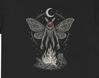 Puede incluir: Camiseta negra con un gráfico detallado de una criatura parecida a una polilla con ojos rojos, alas y una luna sobre una hoguera. El diseño incluye un puente y estrellas, creando una escena mística. La camiseta está hecha de un material suave y cómodo.