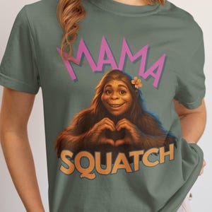May include: Blue Spruce t-shirt featuring a smiling Bigfoot character forming a heart with its hands. The shirt has the words "MAMA SQUATCH" in pink and orange. The text "Blue Spruce" is below the graphic.