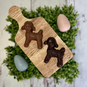 May include: Two chocolate lamb figurines on a wooden cutting board, with a green leafy background and two pastel Easter eggs. The lambs are brown and dark brown, and the eggs are light blue and pink.