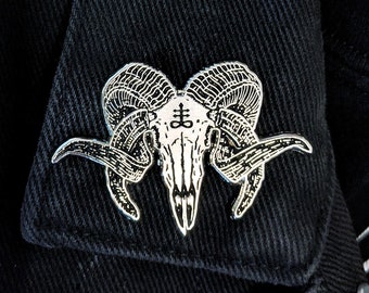 May include: A silver pin with a black and white image of a skull with horns and the symbol of the inverted pentagram on the forehead. The pin is on a black jacket.