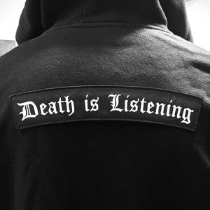 May include: Black hooded sweatshirt with a black patch on the back that says "Death is Listening" in white lettering.