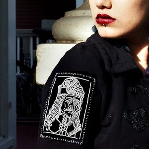 Könnte beinhalten: Eine Frau mit leuchtend blauen Haaren trägt eine schwarze Jacke mit einem weißen gestickten Aufnäher. Der Aufnäher zeigt ein stilisiertes Bild einer Person mit einer Krone und langen Haaren. Die Jacke hat dekorative schwarze Verzierungen.