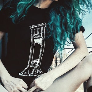 May include: A black t-shirt with a white graphic of a guillotine. The person wearing the shirt has teal blue hair.
