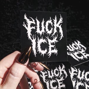 May include: Black sticker featuring the words "FUCK ICE" in a white, gothic font. The sticker is held by a hand with long, gold-coloured nails and copper-coloured rings. Several other stickers are visible.