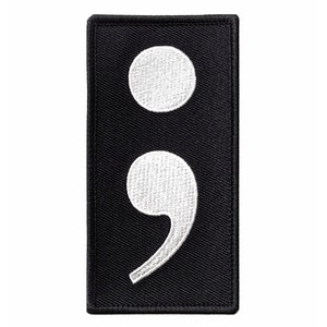 Pode incluir: Patch de tecido preto com um ponto e vírgula bordado em branco.