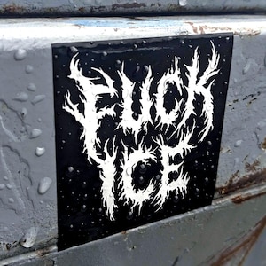 May include: A black square sticker with the words "FUCK ICE" in white, stylized, jagged lettering. The sticker is attached to a weathered, grey metal surface, with water droplets visible on the sticker and surrounding area.