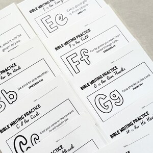 May include: A stack of white Bible writing practice cards. Each card features a letter of the alphabet, a corresponding word, and a Bible verse. The letters are outlined in black, with the words and verses printed in black.