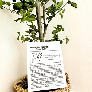 May include: A Bible writing practice sheet with the letter "F" for faith, displayed in a woven basket. The sheet includes the text "For by grace you have been saved through faith" and the scripture reference Ephesians 2:8. A small tree with green leaves is in the background.