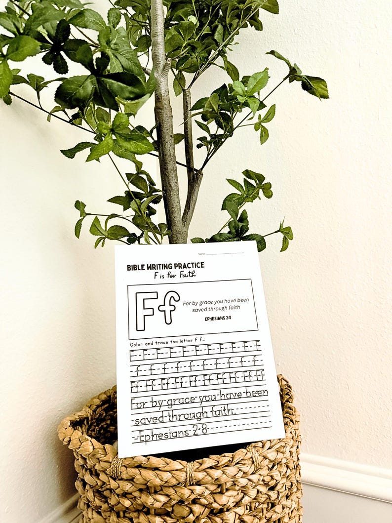 May include: A Bible writing practice sheet with the letter "F" for faith, displayed in a woven basket. The sheet includes the text "For by grace you have been saved through faith" and the scripture reference Ephesians 2:8. A small tree with green leaves is in the background.
