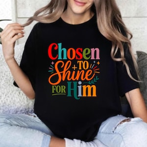 Op de afbeelding: Zwart T-shirt met de tekst "Chosen to Shine for Him" in kleurrijke, gestileerde letters. Het ontwerp bevat rode, oranje, groene en blauwe tinten, met decoratieve elementen. Een casual kledingstuk.