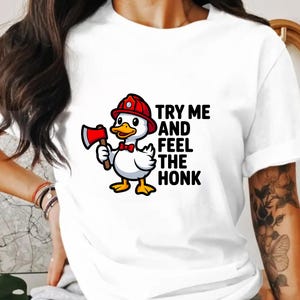 May include: White t-shirt featuring a cartoon duck wearing a red firefighter helmet and holding a red axe. The text "TRY ME AND FEEL THE HONK" is printed on the shirt. The duck has a red bow tie and yellow feet.