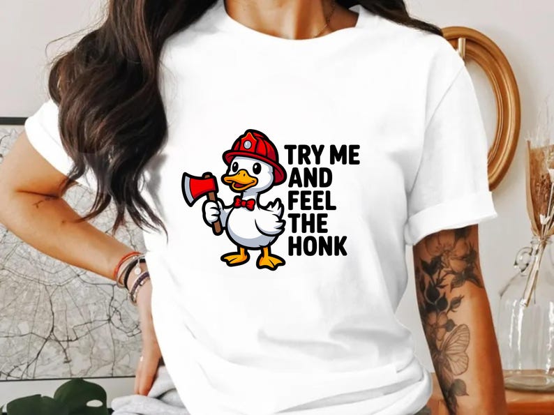 May include: White t-shirt featuring a cartoon duck wearing a red firefighter helmet and holding a red axe. The text "TRY ME AND FEEL THE HONK" is printed on the shirt. The duck has a red bow tie and yellow feet.