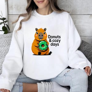 May include: White sweatshirt featuring a cartoon capybara holding a green donut. The capybara is orange and brown with a smiling face. Text on the sweatshirt reads "Donuts & cozy days".