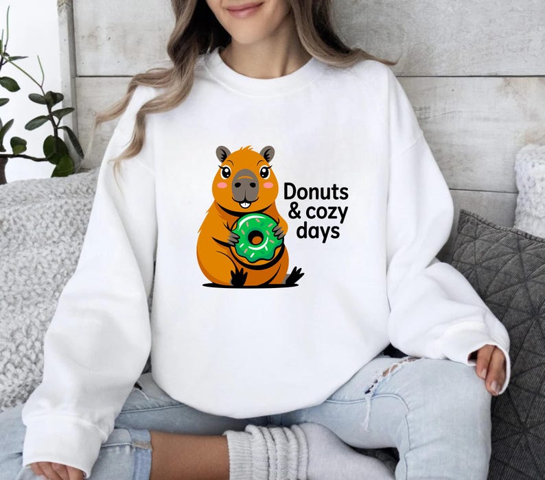 May include: White sweatshirt featuring a cartoon capybara holding a green donut. The capybara is orange and brown with a smiling face. Text on the sweatshirt reads "Donuts & cozy days".